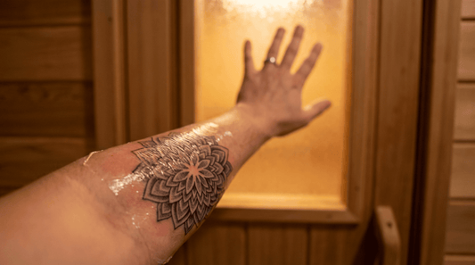 Frisch tätowierter Unterarm mit Schutzfolie vor einer Saunatür als Symbol für die Wartezeit zwischen Tattoo und erstem Saunagang